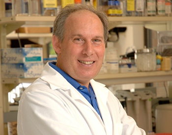 Dennis Drayna, Ph.D., circa 2014, stands in a lab coat in front of one of his lab benches housing various bottles of chemicals.