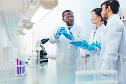 Young male scientist holding flask of liquid in a lab setting, discussing chemical reaction with another young male scientist and young female scientist.