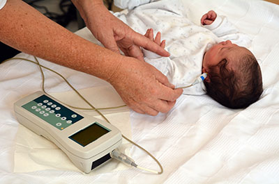 Un bebé recién nacido está acostado boca arriba mientras un adulto inserta una sonda electrónica en su oído. La sonda está conectada a un cable que conduce a un dispositivo portátil que evalúa la audición.