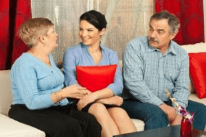 Un hombre mayor está sentado en un sofá junto a una mujer más joven y una mujer mayor. Las dos mujeres hablan mientras el hombre las mira.