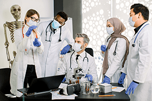 Group of five laboratory scientists in lab coats, masks and gloves, working at lab with test tubes, microscope, flasks, and laptop.