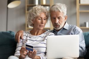 An older woman and man are sitting together on a sofa looking at a laptop computer screen. The woman is holding a credit card.